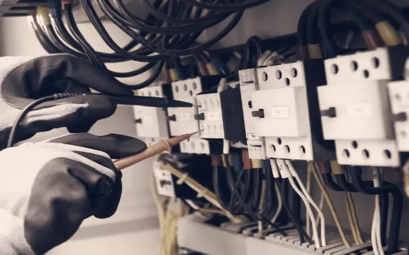 Electrician in insulated gloves testing breakers in an electrical panel with a meter probe