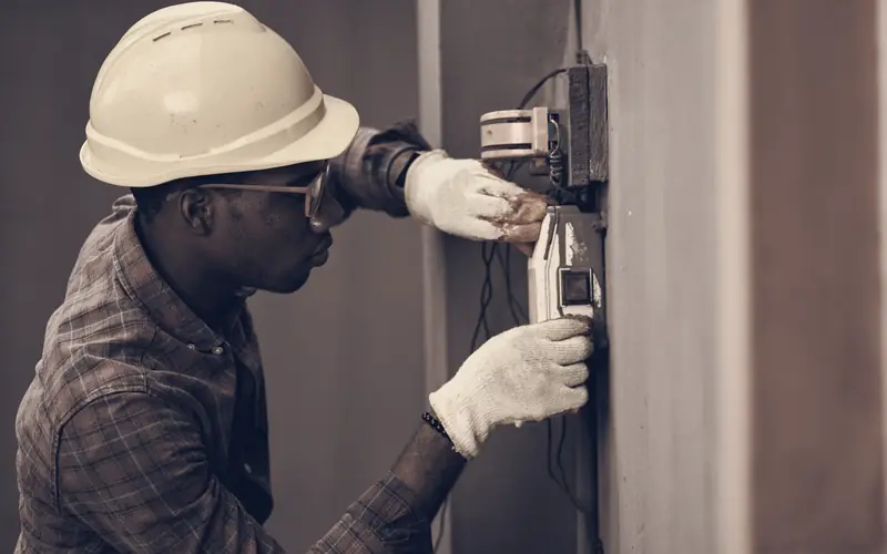 Electrician in a hard hat and safety glasses wiring a wall-mounted meter